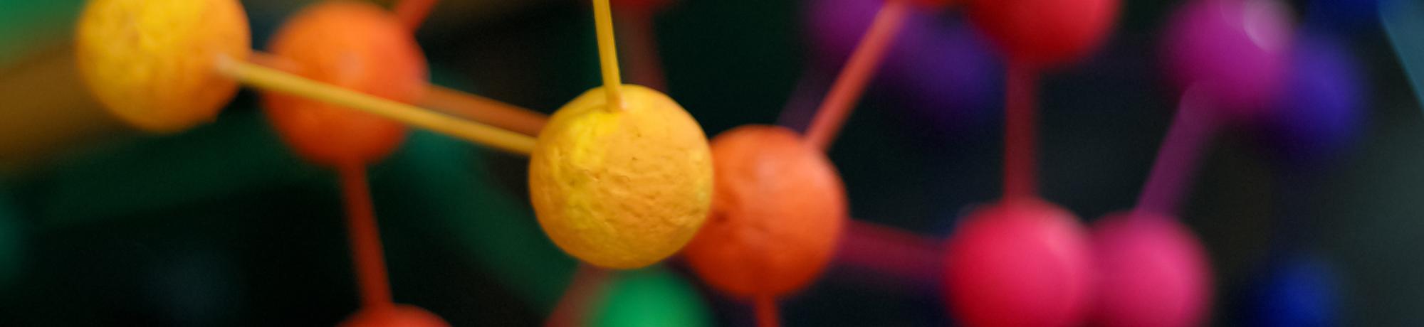 A close-up view of a molecular model with brightly colored spheres representing atoms connected by sticks symbolizing chemical bonds. The spheres in focus are yellow and orange, while the background shows more spheres in colors like purple, blue, and red, slightly out of focus. This model is used to visualize molecular structures and understand the spatial arrangement of atoms within a molecule.